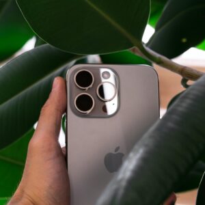 Hand holding a smartphone with triple camera lens against green leaves, showcasing modern design.